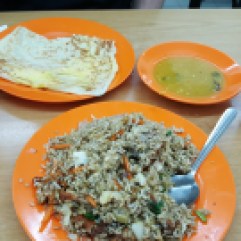 Three plates of food, a small can of CocaCola and a small can of A&W. One plate has a flat bread with egg in it. Another plate is slightly deeper and holds a green sauce. The final plate is a mound of dark colored, fried rice