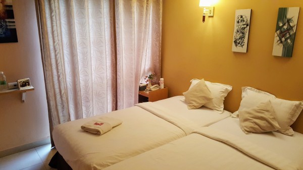 Hotel room with yellow wall and some art, two twin beds pushed together with white sheets, a bedside table with fake flowers and a phone on it, a shelf on the wall with a framed photo and Carbon Monoxide Detector.